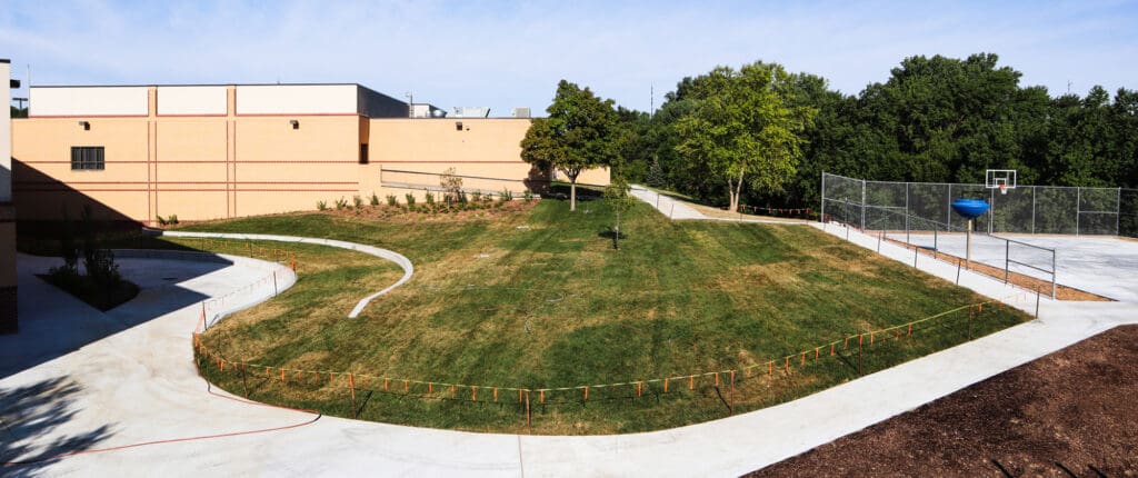 Campus courtyard with curved concrete path, grassy hill, and a fenced basketball court on the right.