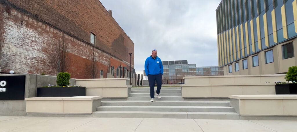 Man in a bright blue jacket descending concrete steps in an urban plaza between a brick wall and a modern glass-and-yellow building behind him.