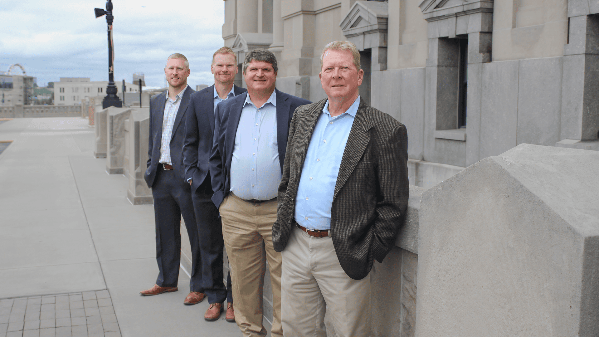 Four men in business attire posing for a photo along a city sidewalk beside a stone building.